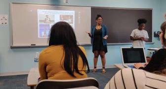 Photo of a woman standing in front of a classroom teaching on a projector.