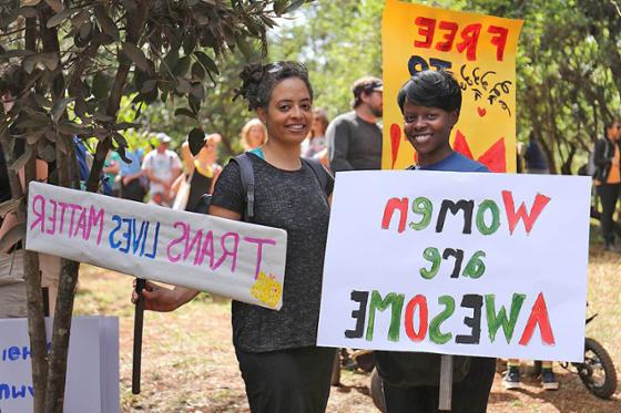 Photo of two young women holding signs reading Women Are Awesome and Trans Lives Matter